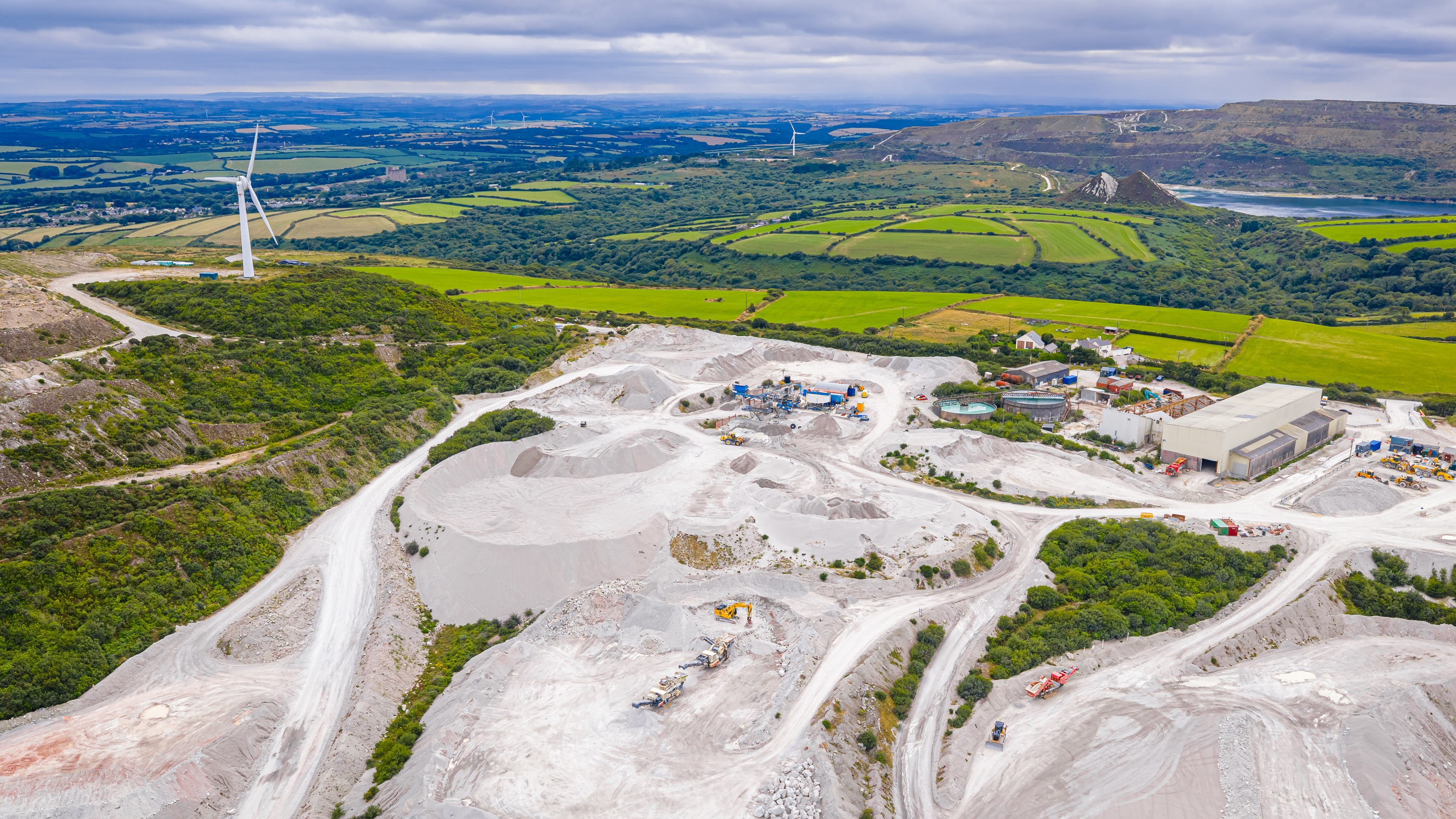 Aerial view of clay mining site in Cornwall with machinery, winding roads, and rural landscape under cloudy skies